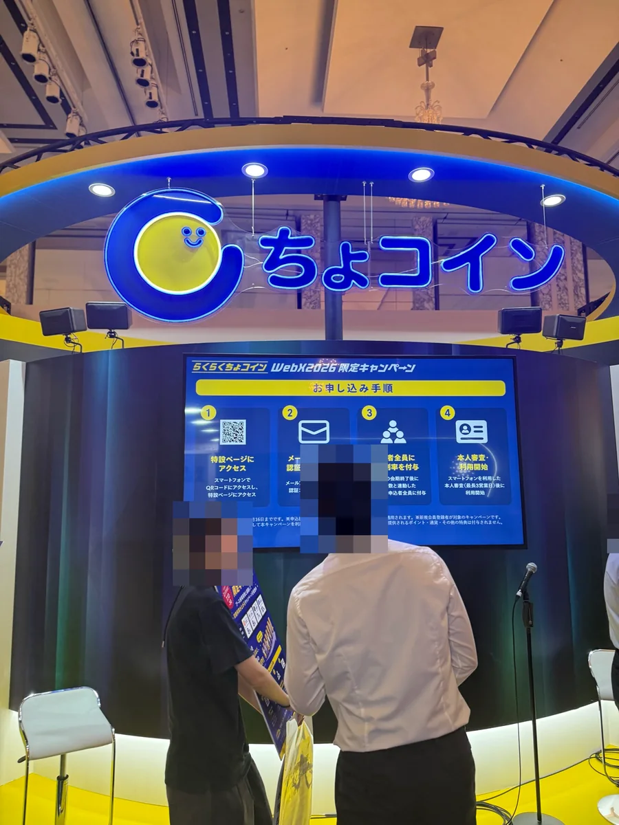WebX 2026のらくらくちょコインのブース。青と黄色のちょコイン看板と当時のキャンペーン案内があり、人物はモザイク加工済み
